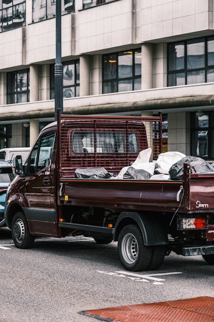 A burgundy flatbed utility vehicle parked on a city street in front of a modern multi-story building with large glass windows and light-colored stone cladding. The truck's open cargo area is filled with various bags, containers, and loose rubbish, suggesting on-site clearance or private waste collection. The vehicle features a protective metal grid behind the cab, and the cargo surface is occupied with items of different materials, including plastic and fabric, in neutral and dark tones. The surrounding environment includes a lamppost and a white van partially visible in the background, with the scene illuminated by natural daylight, creating a neutral, professional setting typical of independent rubbish removal services or alternative waste handling approaches in urban areas. Waste Clearance Brent’s vehicle exemplifies private rubbish collection, often used for non-local authority waste disposal, with a focus on managing domestic or commercial waste outside typical municipal services.