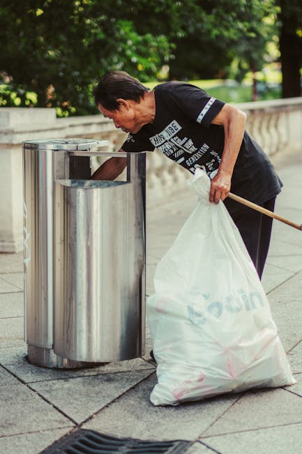 A man with dark hair, wearing a black T-shirt with white text, is seen disposing of a large white plastic bag filled with rubbish into a cylindrical metal trash bin located outdoors on a paved walkway. The bin has a metallic, slightly reflective surface with a rectangular opening at the top, positioned beside a low stone railing. In the background, there is lush green foliage and trees, suggesting a park or public space setting during daylight hours. The man is leaning forward, holding a wooden stick in his right hand, and appears to be carefully ensuring the rubbish is properly deposited. The scene is well-lit with natural light, highlighting the textures of the bag, metal bin, and surrounding environment. This image exemplifies private, independent rubbish collection related to waste clearance services often provided by companies such as Waste Clearance Brent, especially when managing general waste in designated outdoor areas.