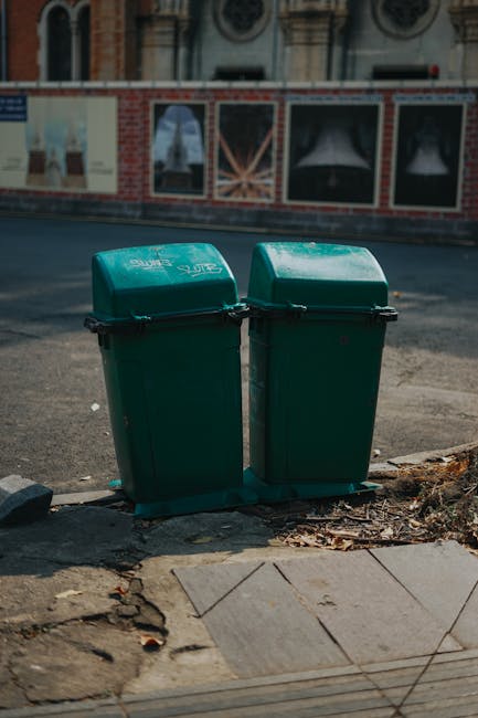 Two green wheeled rubbish bins with black handles and lids, positioned side by side on the edge of a pavement in an urban area. The bins have a smooth plastic surface with a slight sheen, and their lids are closed, showing a subtle curved profile. They are placed on a concrete curb with some scattered debris and fallen leaves nearby. In the background, a brick and plaster building facade with decorative windows and hanging banners or posters is visible, slightly blurred, indicating a city street environment. The scene is lit with natural daylight, casting soft shadows, and the overall setting relates to waste collection or private rubbish disposal, aligning with the topic of alternative waste handling services provided by Waste Clearance Brent in Kilburn.