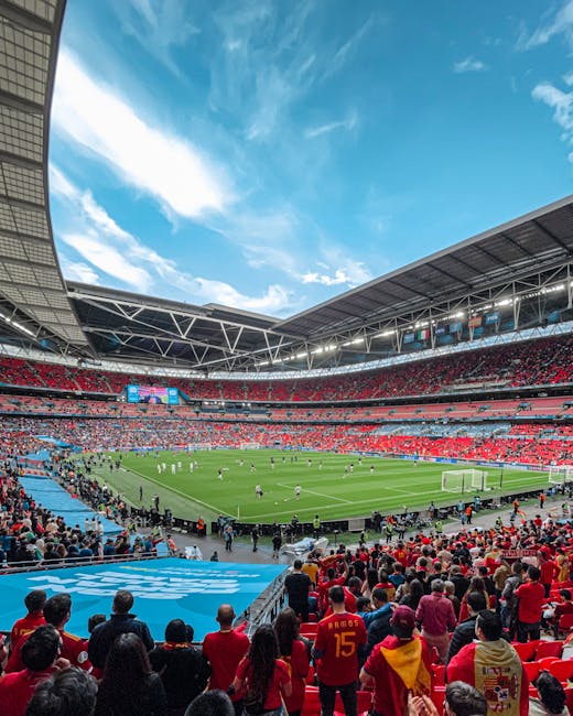 A panoramic view of a large stadium during a football match, with a lush green pitch in the center and players actively engaged in the game. The stadium is filled with spectators, many wearing red clothing, seated in multiple tiers of red and gray seats. The stands are partially covered by a modern, open roof structure with visible metal supports. On one side, a large digital scoreboard displays information, with several banners hanging beneath it. The sky above the stadium is bright blue with scattered white clouds, creating a vibrant atmosphere. In the foreground, a crowd watches the game closely, some standing near the barriers. Surrounding the stadium environment appears clean and well-maintained, with clear pathways and area designated for spectators. Although the image does not explicitly show waste management activities, it reflects typical stadium settings where private waste handling and rubbish clearance are involved in maintaining cleanliness during large events. Waste Clearance Brent provides professional rubbish removal services suitable for such venues, ensuring efficient on-site clearance and alternative waste handling solutions.