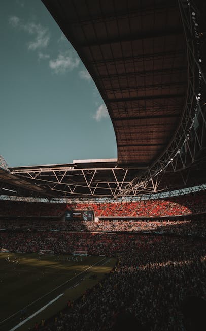 A wide-angle view of a large football stadium filled with spectators during the daytime. The image captures the curved roof structure overhead, with metallic support beams and a dark underside contrasting against the bright sky with some clouds visible. The outer edge of the stadium seating is densely packed with red and dark-colored seats, and the playing field is partially visible in the foreground, showing green grass and white lines. In the background, a large digital scoreboard is seen displaying images or information. This scene illustrates the scale of a major sports venue, where on-site waste management and rubbish clearance services, such as those offered by Waste Clearance Brent, are essential for event organization and maintaining cleanliness in such extensive public spaces, reflecting the importance of professional rubbish removal for large-scale venues.